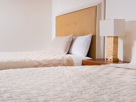 Habitación de hotel con dos camas, cabecero de madera, mesita de noche con lámpara y otra mesita.