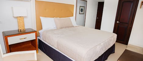 A hotel room with a large bed, a bedside table with a lamp, a wooden bench, and a door.