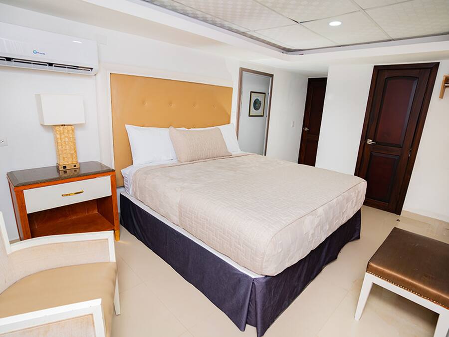 Een hotelkamer met een groot bed, een nachtkastje met een lamp, een houten bankje en een deur.