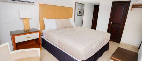 A hotel room with a bed, a desk, a chair, a small stool, and a door.