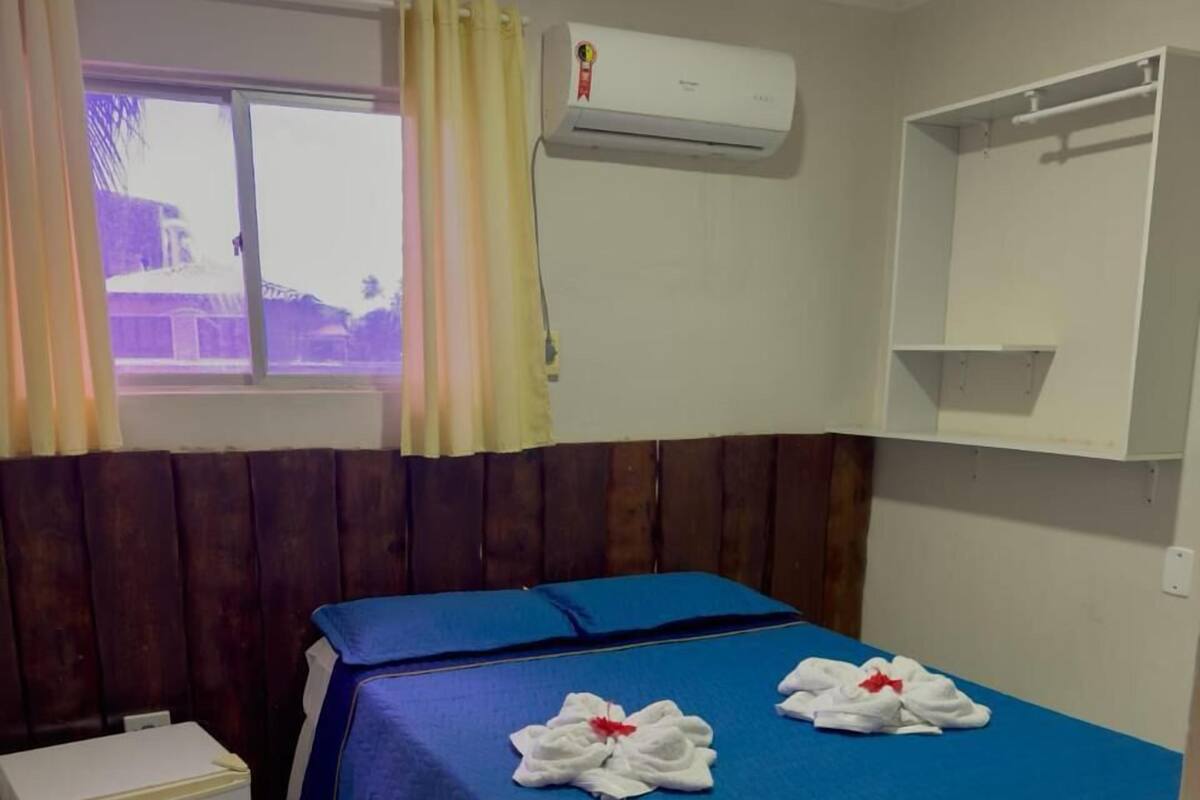 Hotelkamer met een bed, een kleine witte koelkast, een airconditioner, een raam met gordijnen en een wandplank.