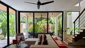 Ein modernes Wohnzimmer mit Sofa, Couchtisch und Essbereich. Eine Treppe befindet sich rechts, und ein großes Fenster bietet Blick auf einen Garten.