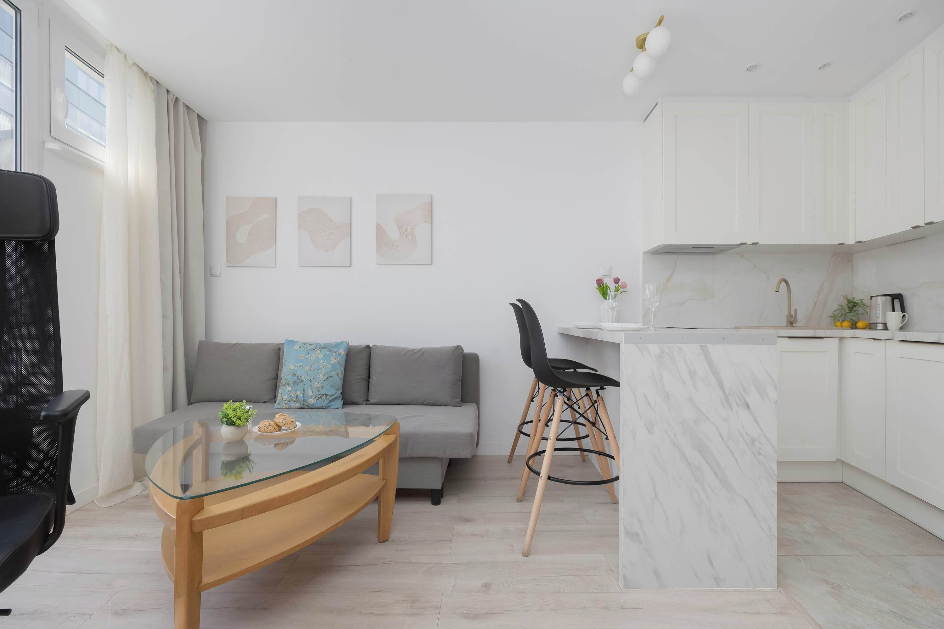 Una sala de estar moderna con una mesa de centro de vidrio, un sofá gris con cojines, una silla negra y una cocina con armarios blancos y una isla de mármol.