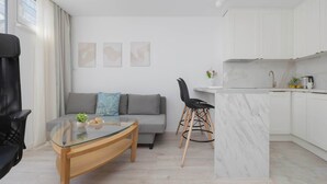 A modern living room with a glass-top coffee table, a grey sofa with cushions, a black chair, and a kitchen area with white cabinets and a marble island.