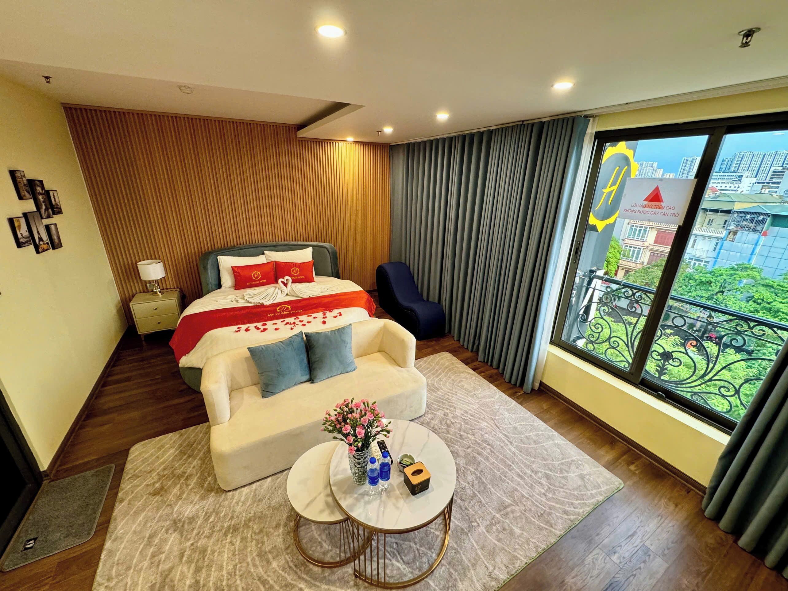 A modern hotel room with a large bed, a sofa, a small round table, and a view of the city through large windows.