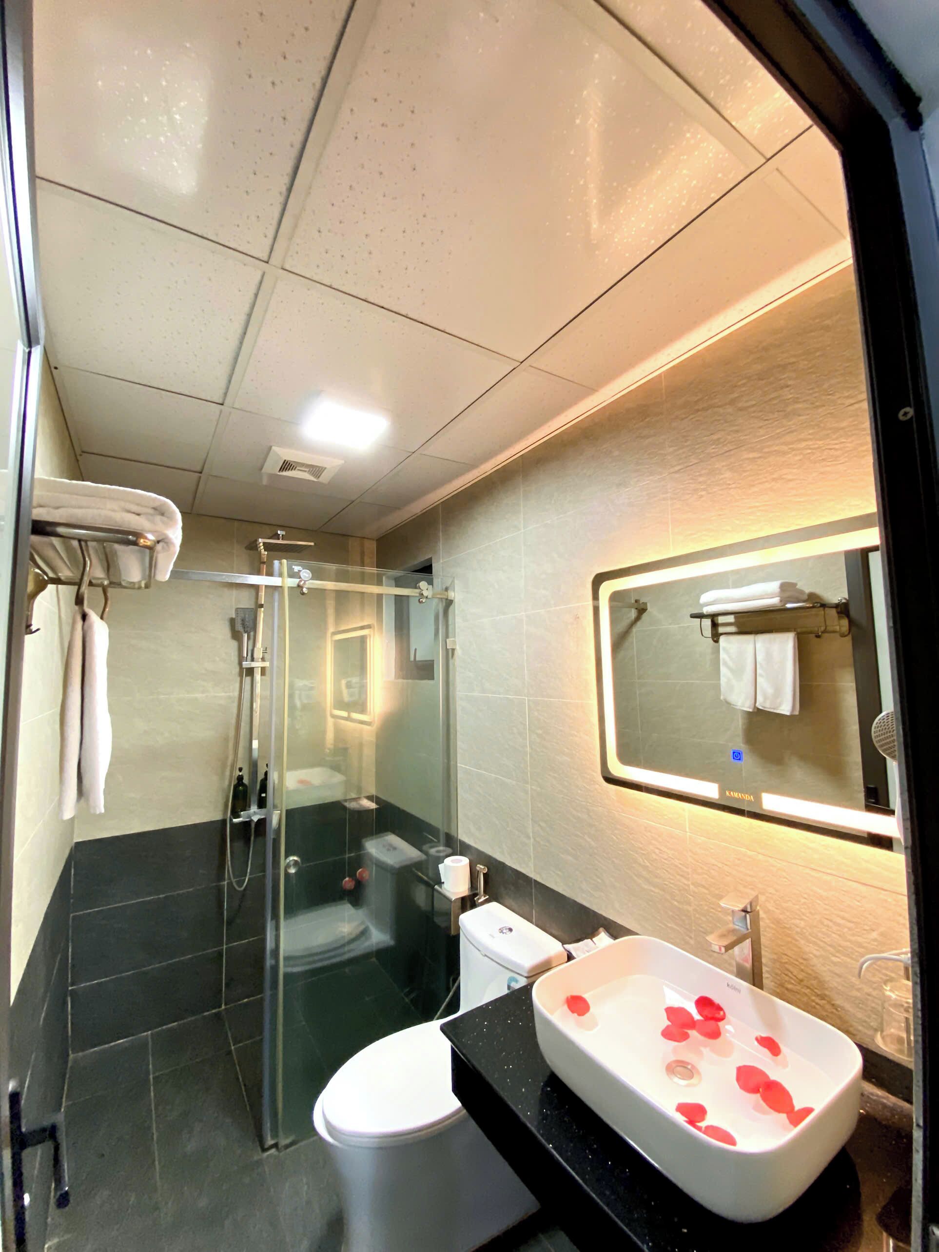 Un baño moderno con ducha de vidrio, un lavamanos blanco adornado con pétalos de rosa, un inodoro y un perchero para toallas.