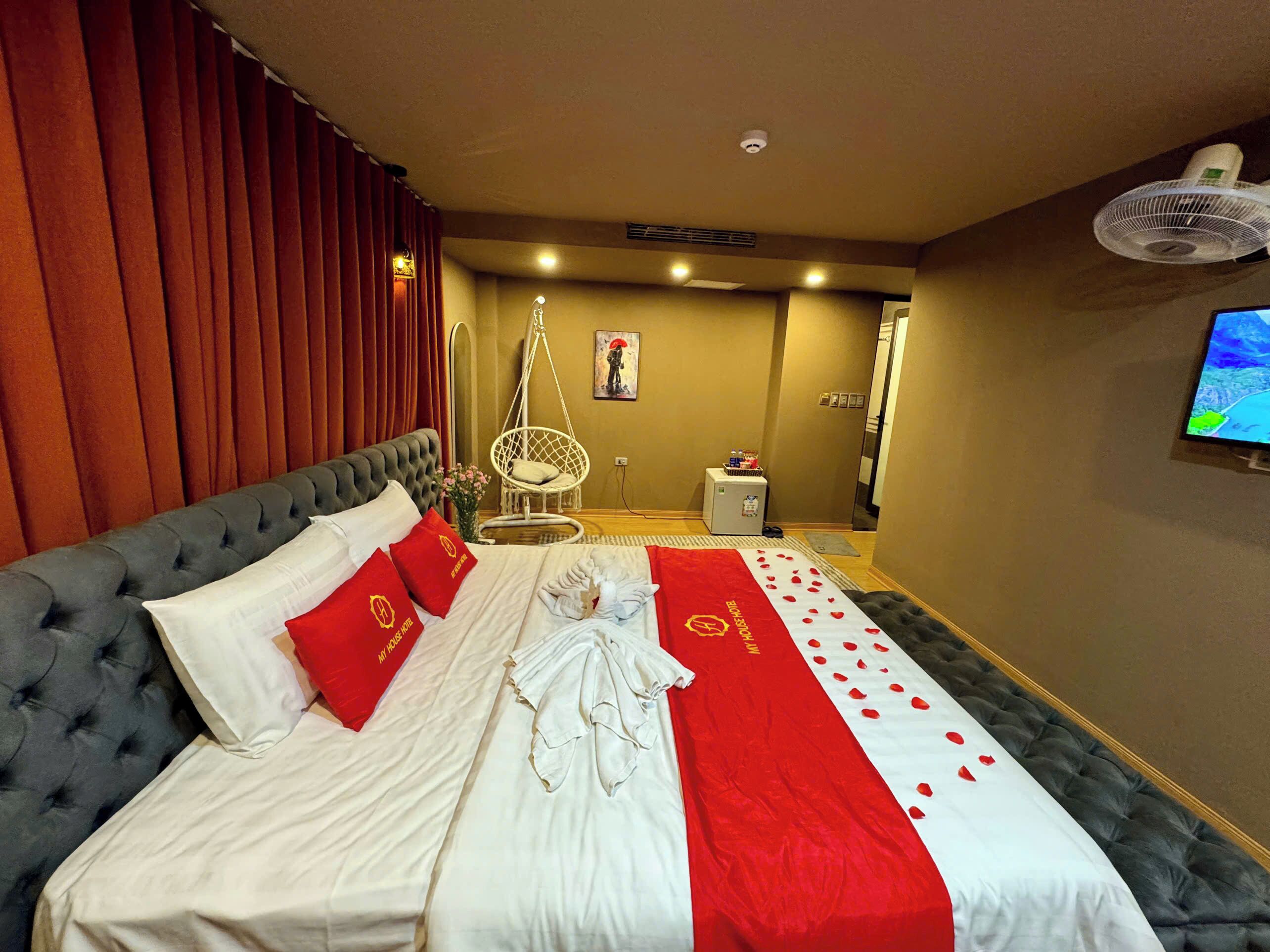 Ein Hotelzimmer mit einem großen Bett, rot-weißer Bettwäsche, einem Hängekorbsessel, einem kleinen Tisch mit Ventilator und einem an der Wand befestigten Fernseher.