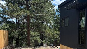 A rustic outdoor area with a wooden deck, stone pathway, and a large pine tree. - Luxurious, Riverside Chalet! Romantic Mountain Views (Johnson Village)