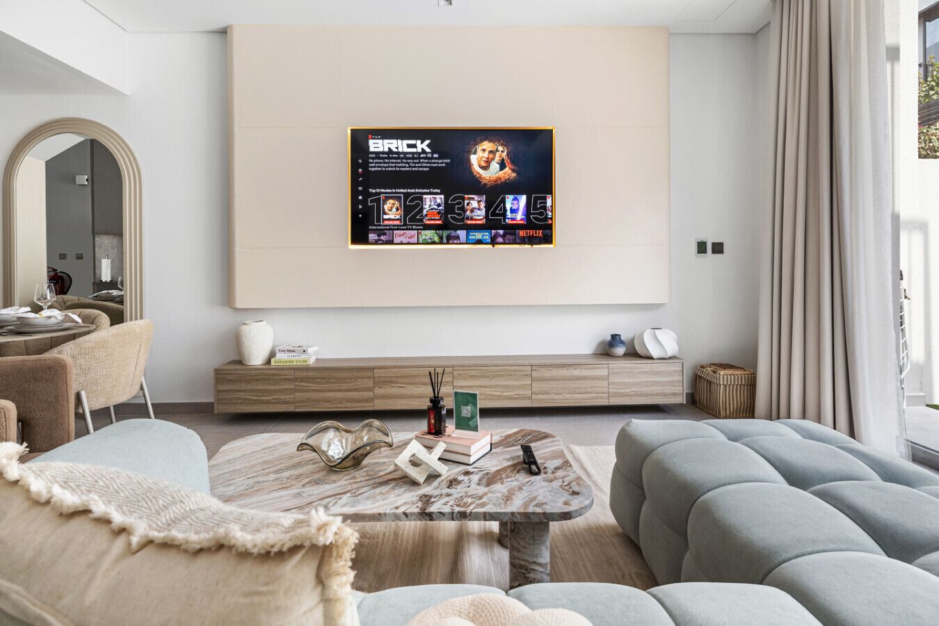 Un soggiorno moderno con una grande TV a schermo piatto montata a parete, un divano, un tavolino da caffè con libri e un vaso, e un mobile contenitore con oggetti decorativi.