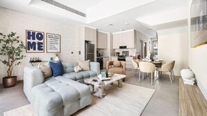 A modern living room with a sectional sofa, a coffee table, and a dining area with chairs. There is a kitchen in the background with cabinets and appliances.