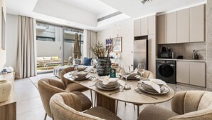 A modern dining area with a round table set for six, surrounded by beige chairs. A large vase with branches is placed on the table.