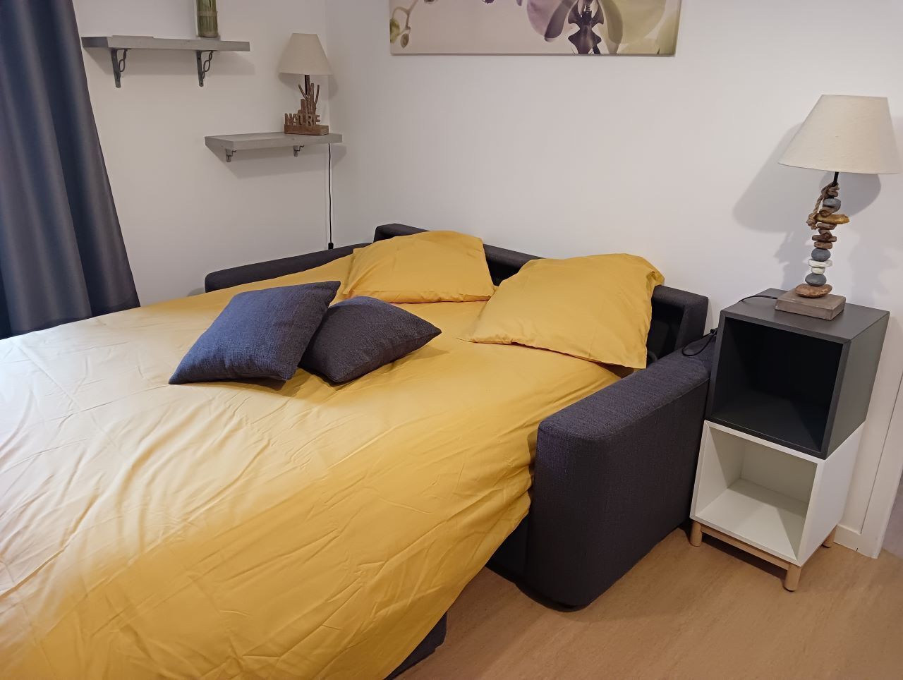 A bed with a yellow bedspread and two pillows, a gray bench, a nightstand with a lamp, and a wall with a painting.