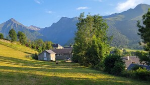 Une maison avec un toit en pente, entourée d’une pelouse, d’arbres et de montagnes en arrière-plan.