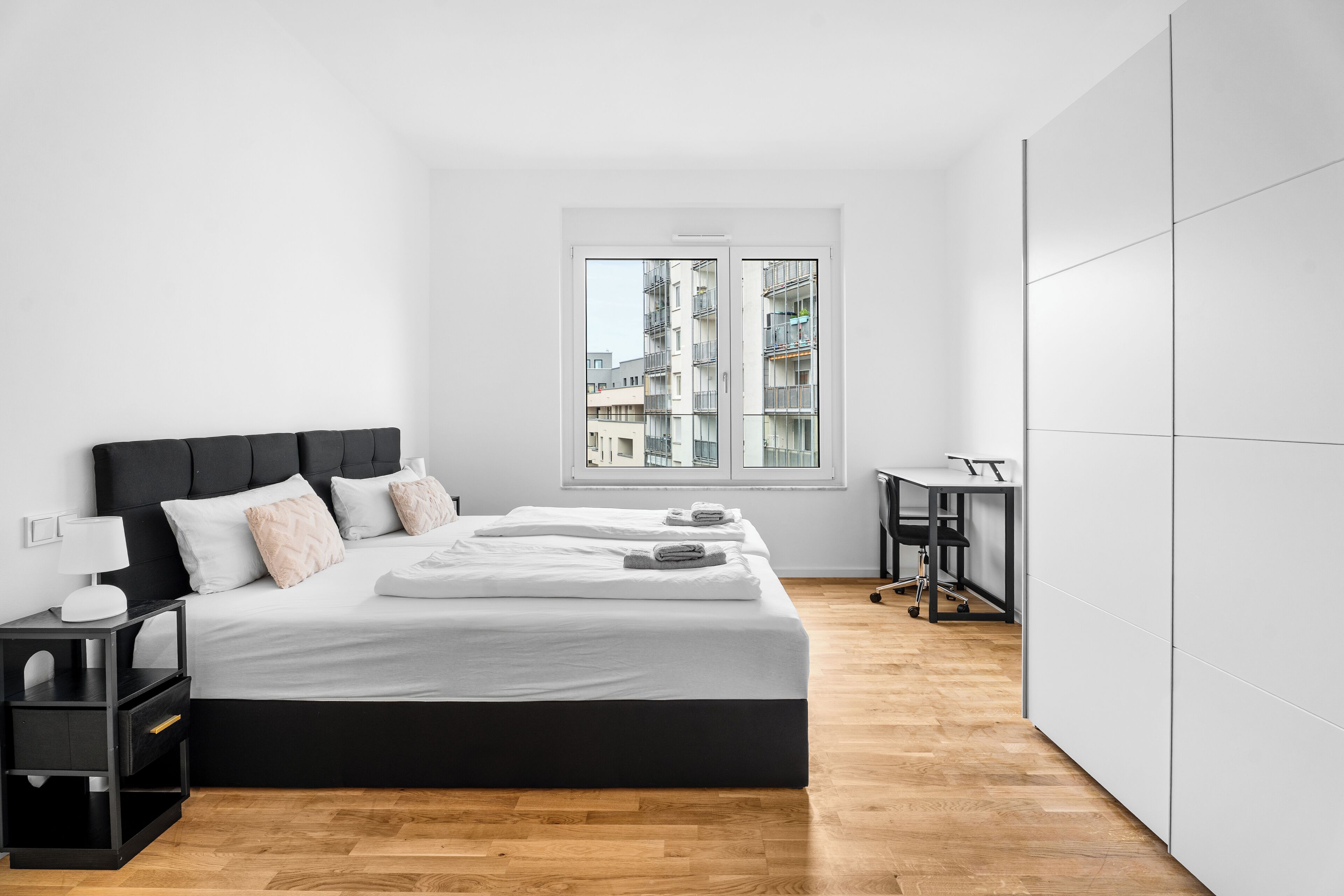 Ein modernes Schlafzimmer mit einem großen Bett, einem Schreibtisch und Blick auf Gebäude durch ein Fenster.