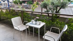 Ein Balkon mit zwei weißen Metallstühlen und einem kleinen Tisch, umgeben von Grünpflanzen und einem Metallzaun.