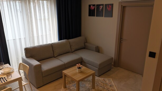 A modern living room with a grey sectional sofa, a small wooden table with a flower vase, a dining area with a table and chairs, and a door with a wall-mounted light switch. - KONYAALTI PLAJINA YAKIN,ŞIK VE YENİ 1+1 SUİT DAİRELER (Antalya)