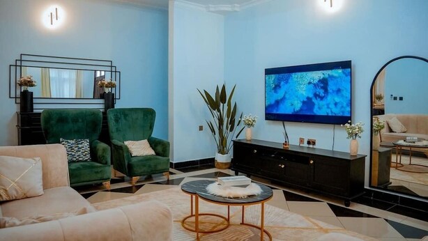 Een moderne woonkamer met een grote flatscreen-tv, groene fauteuils, een beige bank, een zwarte entertainmentkast en een decoratieve spiegel.