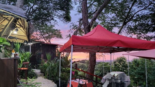 An outdoor dining area with a red canopy, a barbecue grill, and a rustic wooden structure. - Cozy wooden cabin with every comfort and a view of the nearby golf course. (Jicaral)