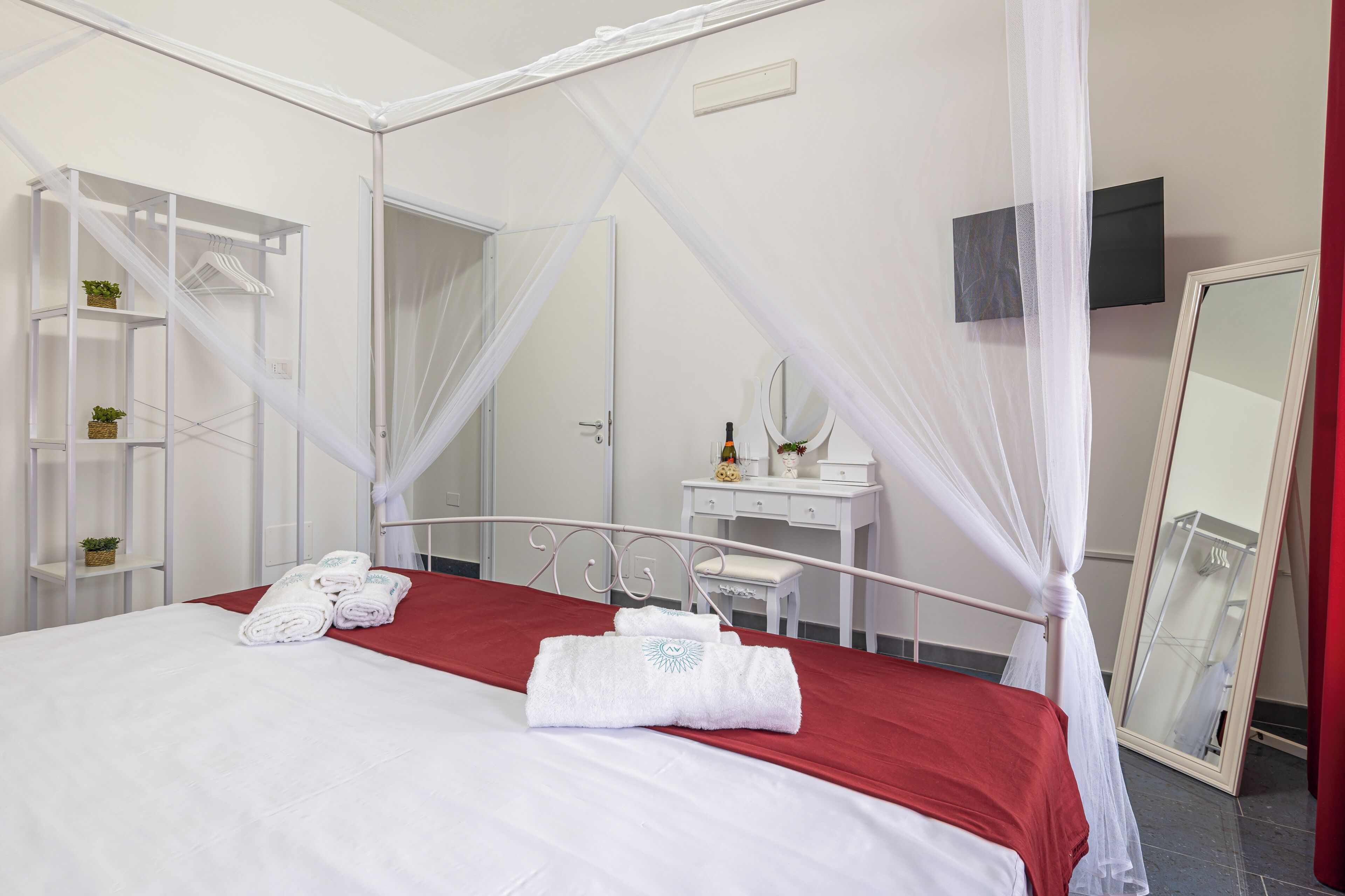 A hotel room with a bed, a vanity with a mirror, a red bedspread, and a white canopy.