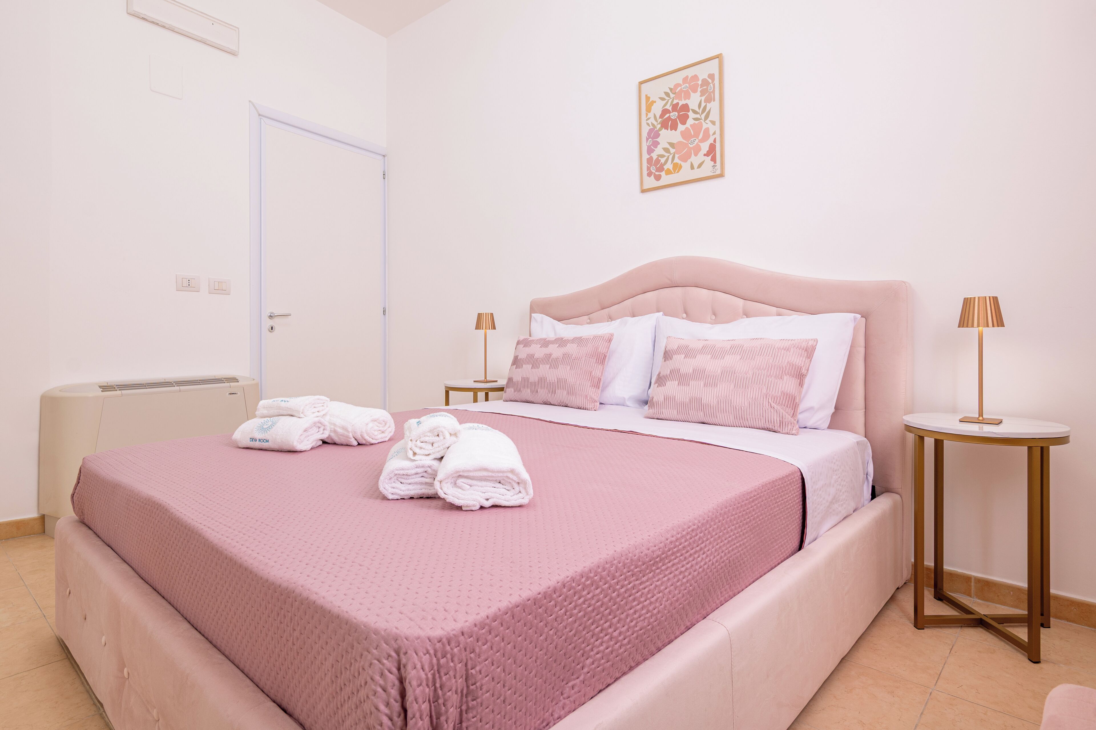 A neatly made bed with pink bedding and rolled towels, a bedside table with a lamp, and a wall-mounted picture.