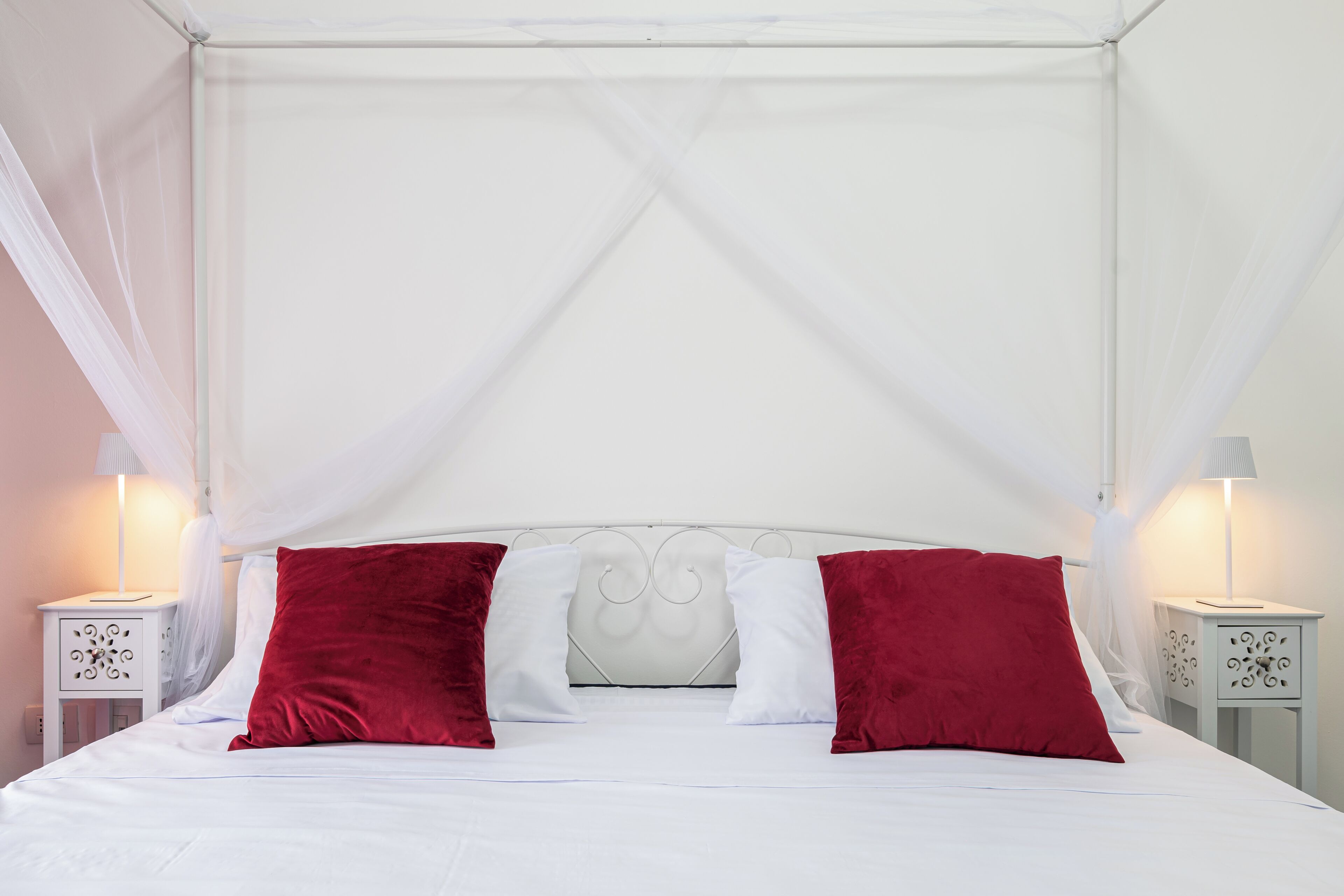 A four-poster canopy bed with white bedding and red pillows, two bedside tables with lamps, and a white wall.