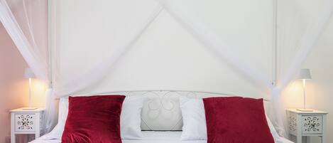 A four-poster canopy bed with white bedding and red pillows, two bedside tables with lamps, and a white wall.