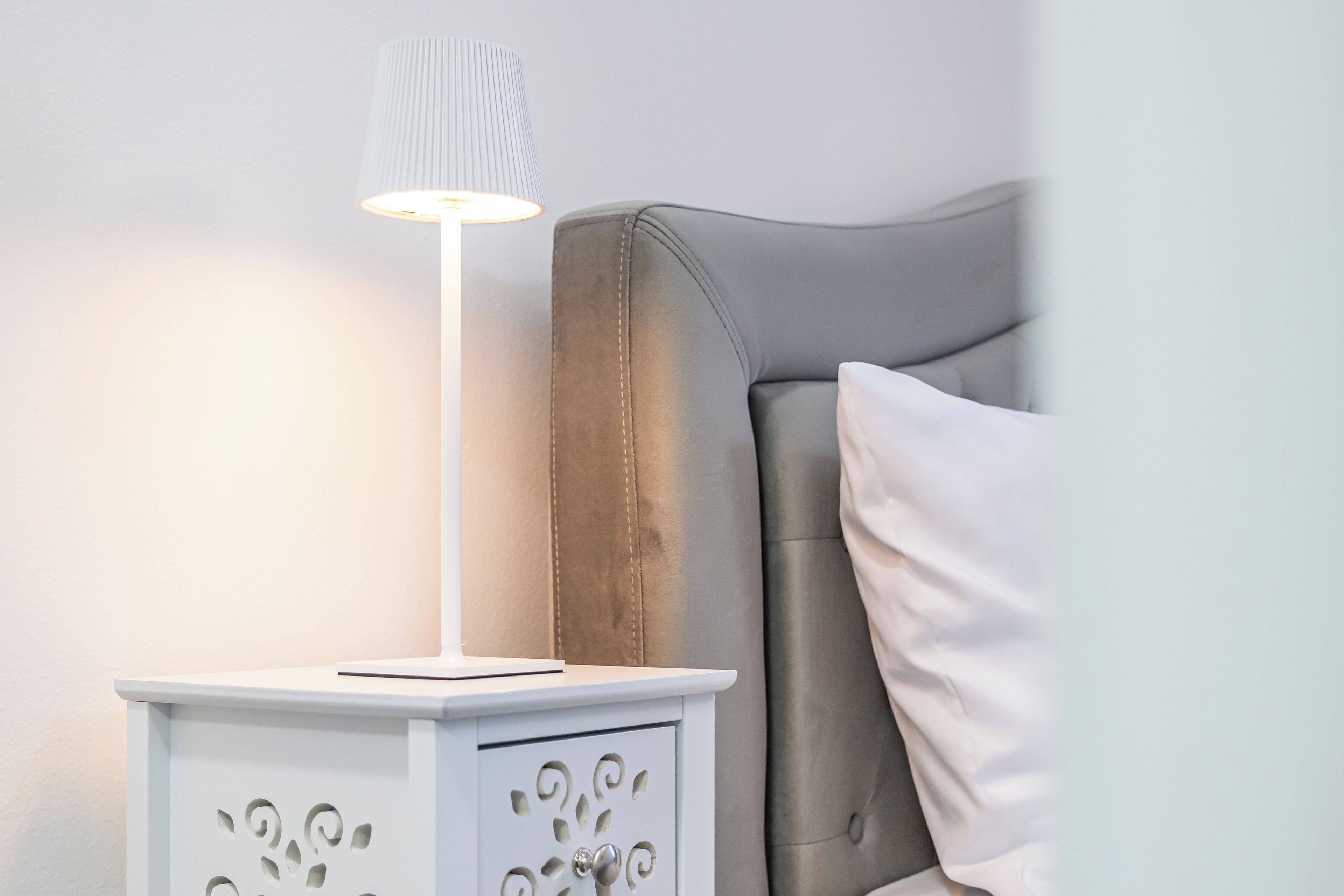 A bedside table with a lamp, a bed with a grey headboard, and a white pillow.
