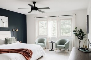 A bedroom with a bed, two chairs, a small table, a ceiling fan, and a window with curtains.