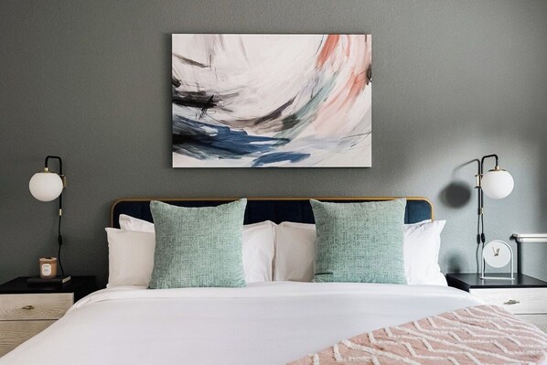 Un dormitorio con cama, almohadas, un cuadro abstracto colgado en la pared, lámparas de noche y una mesita de noche con un reloj.