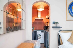 A compact room with a washing machine, a bed, a small table, and a kitchenette.