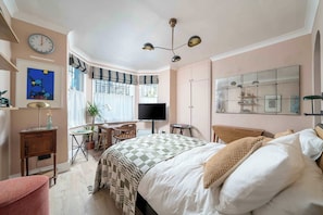 A bedroom with a bed, a desk, a chair, a television, and a large window with curtains. - Charming studio flat near Notting Hill (London)