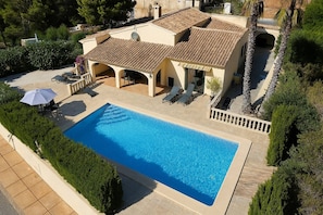 A large swimming pool with a tiled deck, surrounded by a covered patio area with lounge chairs and umbrellas.