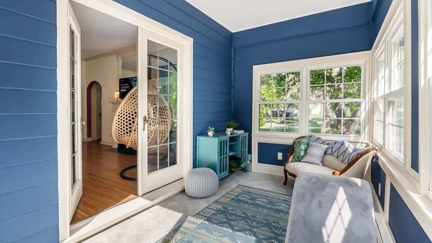Una habitación con pared exterior azul, un ventanal grande, una silla de mimbre, un sofá azul y una pequeña mesa con adornos.