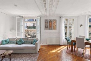 A room with a sofa, dining table, chairs, a radiator, and a window with a view of buildings.