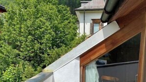 A balcony with a view of a forested hillside, a white wall, and a window with a curtain.