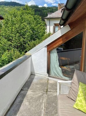 A balcony with a view of a forested hillside, a white wall, and a window with a curtain.