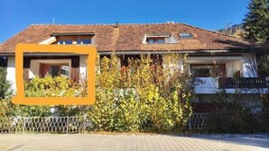 Une maison à deux étages, avec un toit recouvert de tuiles, un balcon bien visible au deuxième étage et un jardin où les plantes ont poussé de manière luxuriante à l’avant.