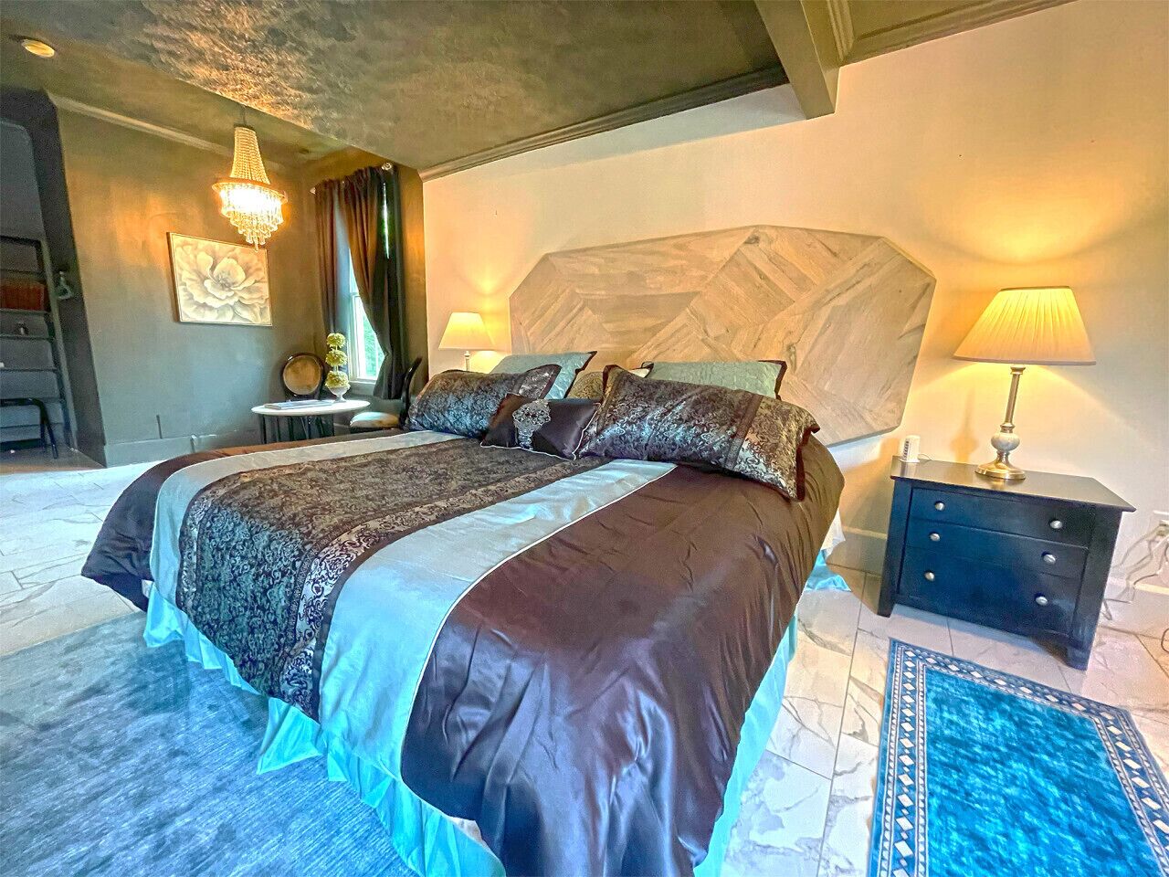 Un grand lit avec une couvre-lit à motifs et plusieurs oreillers, une table de chevet avec une lampe, un élément décoratif mural et un tapis bleu à motifs.