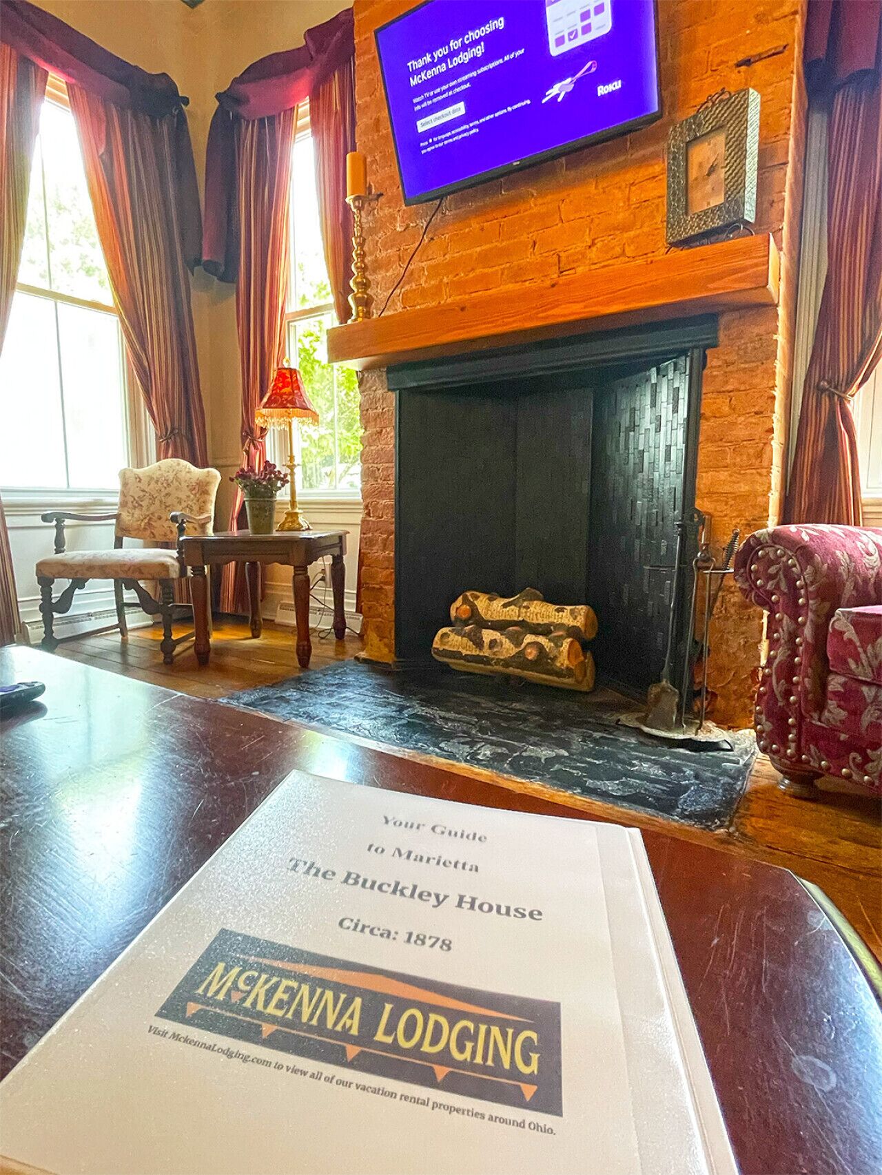 A room with a fireplace, a television screen, a table with a guidebook, and a window with curtains.