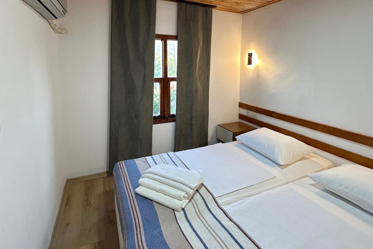A room with a wooden ceiling, a bed with white bedding, a window with curtains, and a wall-mounted light.