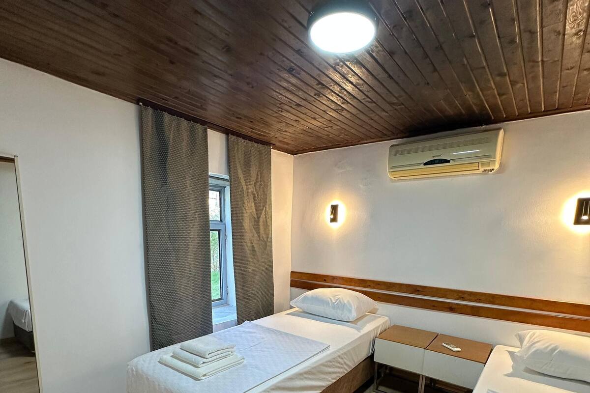 A hotel room with two single beds, a wooden ceiling, an air conditioning unit, and a window with curtains.