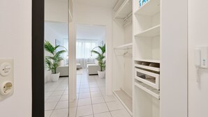 Ein heller, moderner Raum mit weißen Wänden, gefliestem Boden und einem weißen Kleiderschrank mit Einlegeböden. Im Hintergrund stehen zwei Kübelpflanzen und ein weißes Sofa.