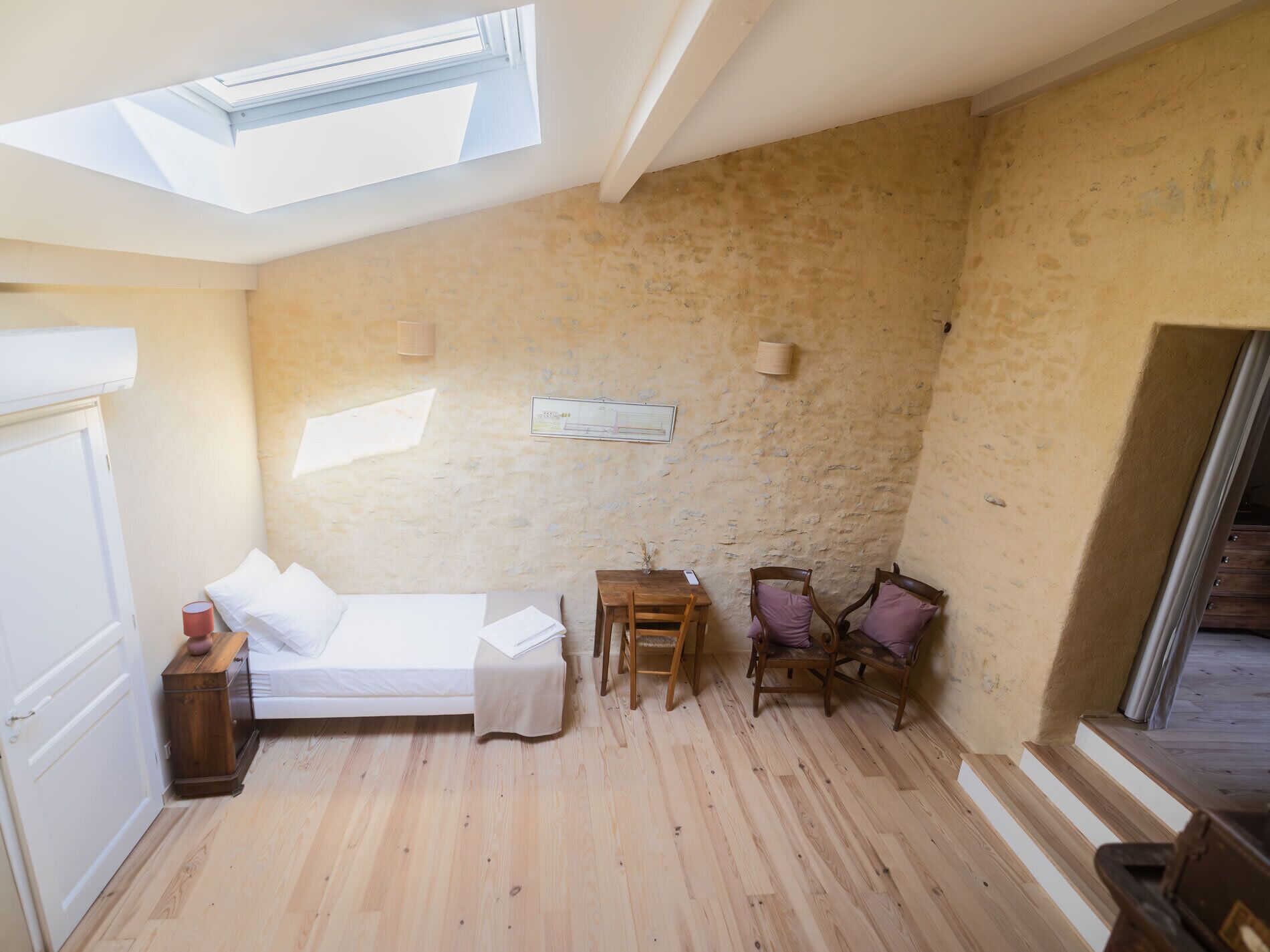 Une chambre avec un puits de lumière, un lit, une petite table, deux chaises et un banc en bois.