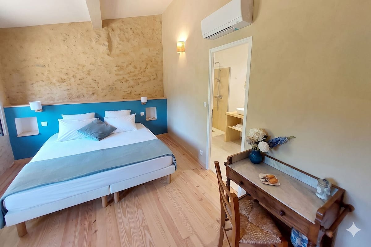 Habitación de hotel con cama, escritorio con espejo, mesita con un florero y un plato, y unidad de aire acondicionado.