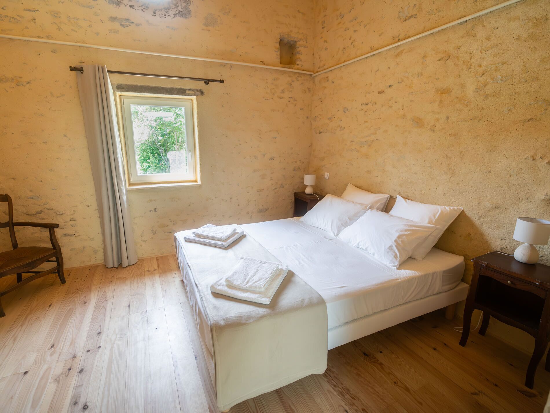 Un dormitorio con suelo de madera, una cama con sábanas blancas, una mesita de noche, una silla y una ventana con cortinas.