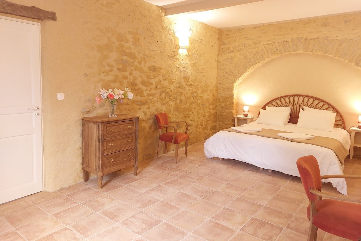 Un dormitorio con pared de piedra, cómoda de madera, cama con ropa blanca y una silla roja.