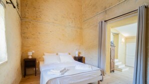 Un dormitorio con piso de madera, una cama con ropa blanca, dos mesitas de noche con lámparas y vista a un pasillo con una puerta.