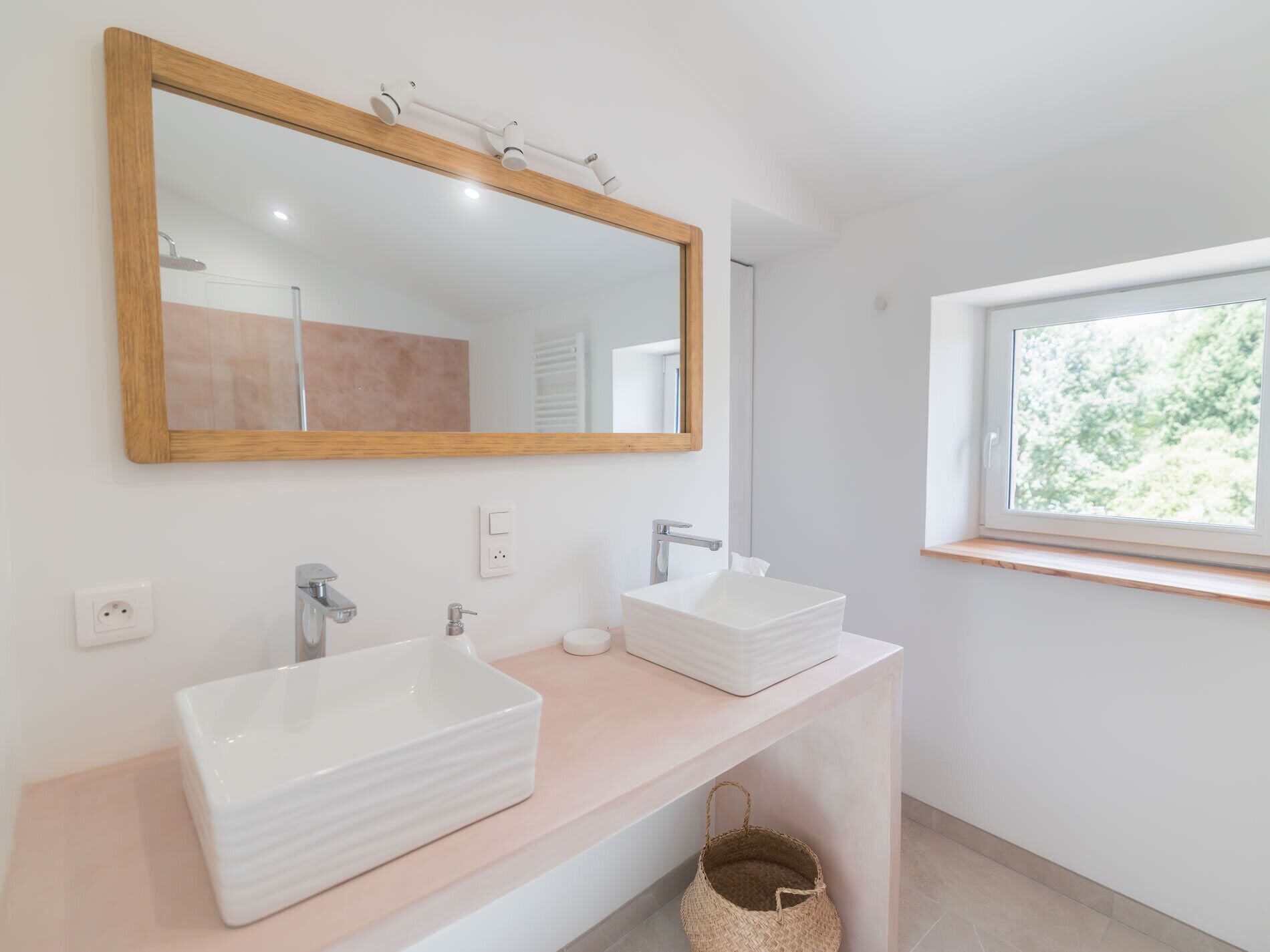 A modern bathroom with two white sinks, a large mirror with a wooden frame, and a window providing natural light.