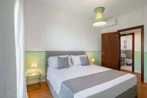 Una cama bien tendida con un edredón gris, dos lámparas de noche y un aplique verde en un cuarto con una puerta de madera y un baño visible a través de una puerta abierta.
