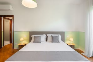 A hotel room with a large bed, two bedside tables with lamps, a ceiling light, and a door leading to another room.